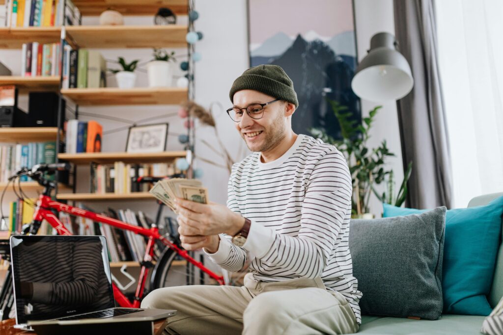 Smiling man counting money while sitting on a sofa with a laptop in a cozy living room setting.
