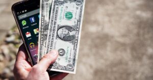 Close-up of a hand holding US dollar bills and a smartphone outdoors, showcasing financial technology.