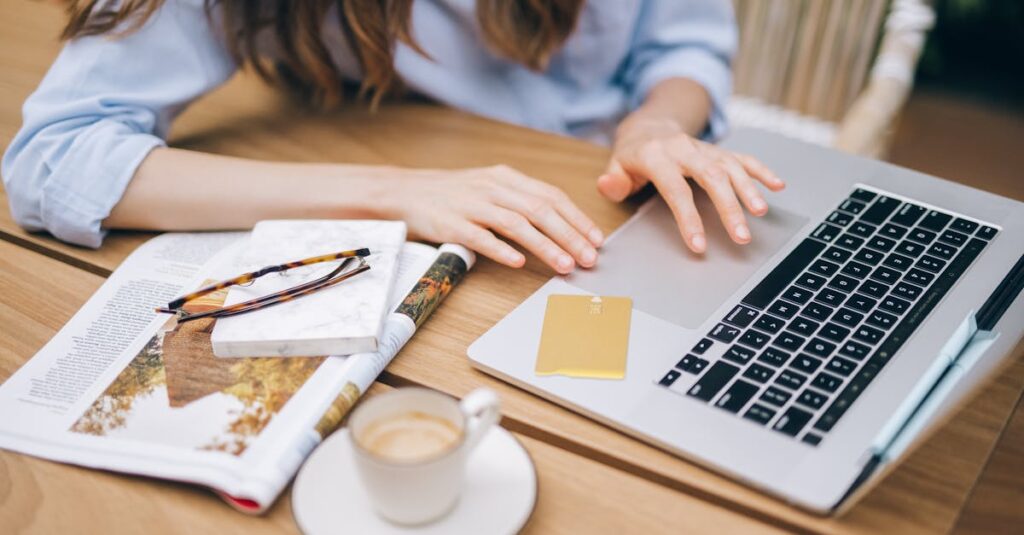 Woman working on a laptop with coffee and a magazine indoors, featuring a casual setting.